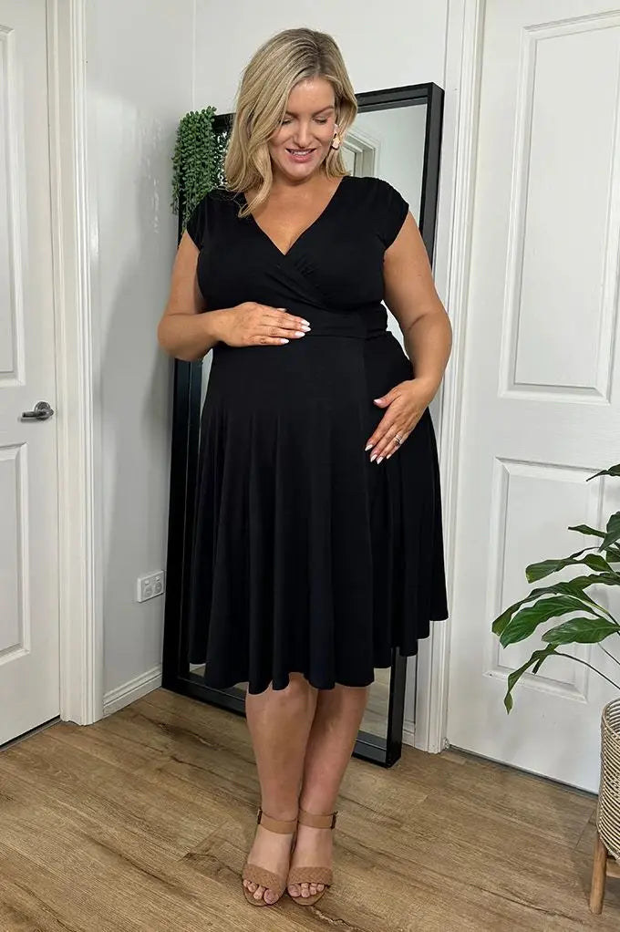 pregnant Woman wearing a black dress standing in a room with white walls and wooden flooring.